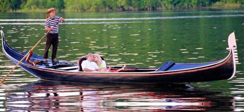 A gondolier steers a black gondola with a couple enjoying a romantic moment on a serene lake.