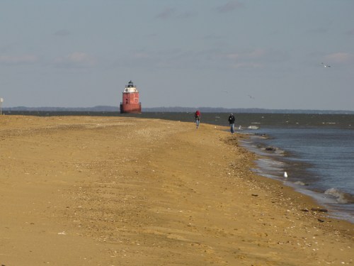 A sandy beach with two people walking, and a distant red lighthouse on the water's edge under a clear sky.