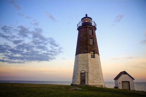 A tall lighthouse stands by the shore at sunset, with a small building nearby and clouds in the sky.