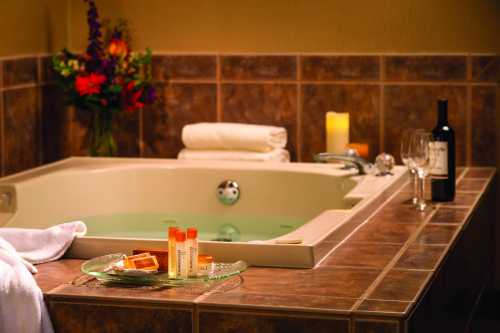 A relaxing bathroom scene featuring a hot tub, candles, wine, and spa products on a tiled countertop.