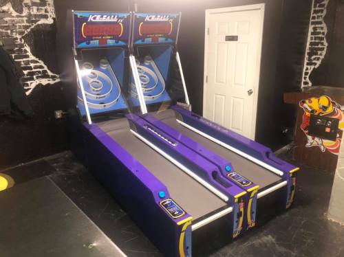 Two purple skee-ball machines side by side in a game room, with a door and brick wall in the background.