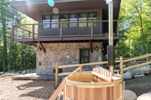 A modern cabin with a stone base and wooden balcony, surrounded by trees, featuring a wooden hot tub in the foreground.