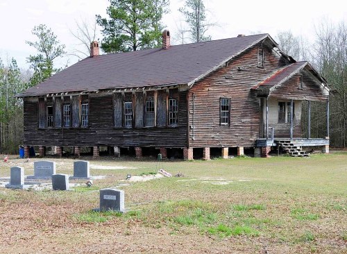 An old, weathered wooden house stands near a small graveyard with several headstones in a grassy area.