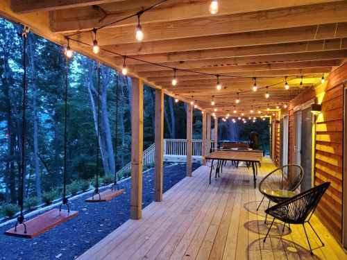 A cozy porch with string lights, wooden flooring, and swings, surrounded by trees in a serene setting.