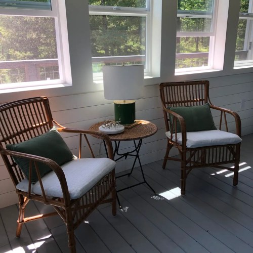 Two wicker chairs with cushions and a small table with a lamp, set in a bright, airy room with large windows.