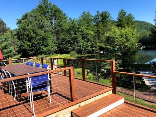 A wooden deck with a dining table and chairs overlooks a lush green landscape and a calm body of water.
