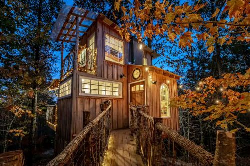 A charming treehouse with wooden walls, large windows, and a bridge, surrounded by autumn foliage at dusk.
