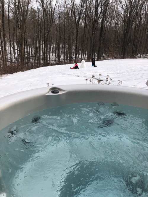 A hot tub in the foreground with snowy landscape and two people building a snowman in the background.