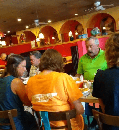 A group of people dining at a restaurant, seated around a table with food and drinks. Brightly colored decor in the background.