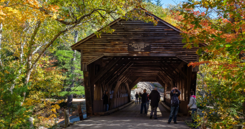 A wooden covered bridge surrounded by colorful autumn foliage, with people walking and taking photos inside.