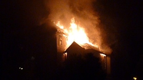 A house engulfed in flames at night, with smoke billowing from the roof and bright fire illuminating the scene.