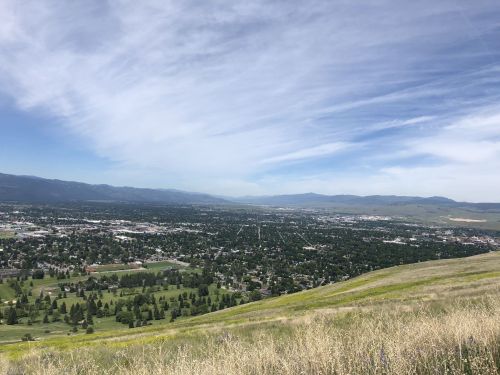 A panoramic view of a valley with a city, green fields, and distant mountains under a partly cloudy sky.