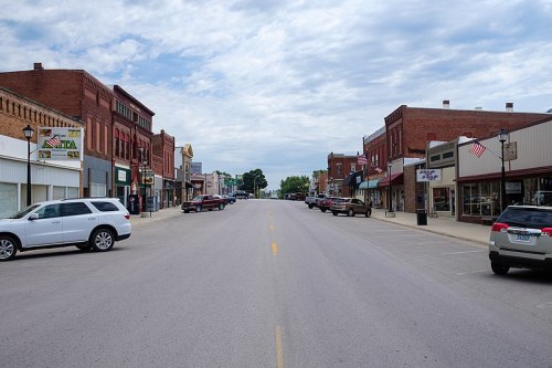 A quiet main street lined with brick buildings, shops, and parked cars under a cloudy sky.