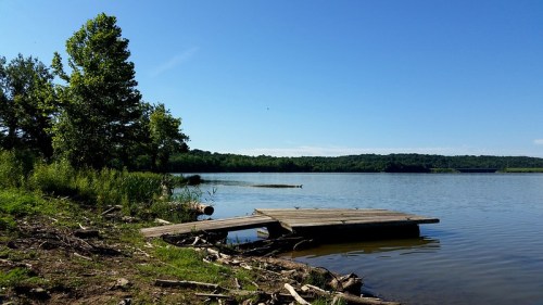 A serene lake scene with a wooden dock, surrounded by greenery and clear blue skies.