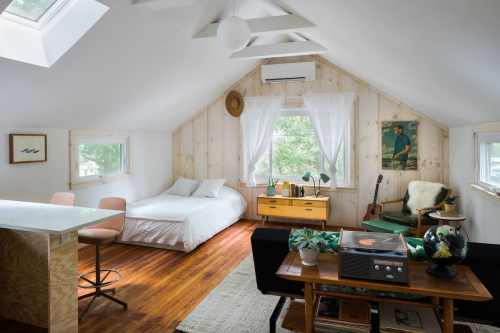 Cozy attic bedroom with a bed, seating area, and natural light from windows, featuring wooden walls and stylish decor.