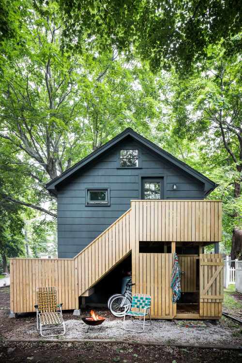 A modern two-story cabin with wooden stairs, surrounded by trees, featuring outdoor seating and a fire pit.
