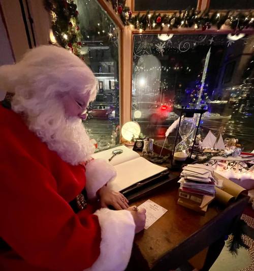 Santa Claus in a cozy room, writing in a book by a window decorated for the holidays, with festive lights outside.