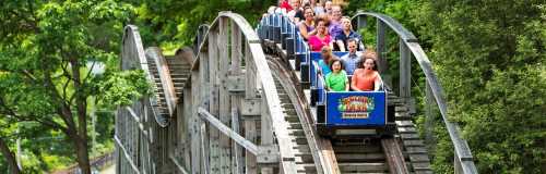 A wooden roller coaster with excited riders ascending a hill surrounded by lush green trees.