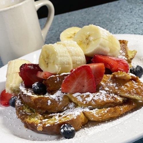 A plate of French toast topped with banana slices, strawberries, blueberries, and powdered sugar, with a coffee mug in the background.