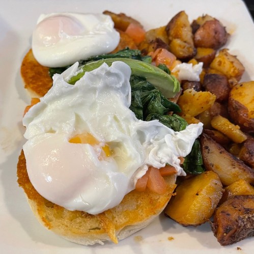 A plate featuring two poached eggs on a biscuit with spinach and tomatoes, alongside crispy roasted potatoes.