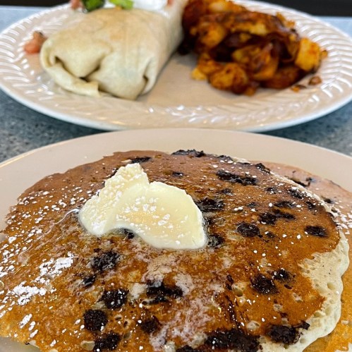 A plate of pancakes topped with butter, alongside a burrito and seasoned potatoes in the background.