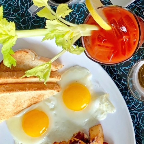 A plate with two sunny-side-up eggs, toast, and a drink garnished with celery and lemon on a patterned tablecloth.