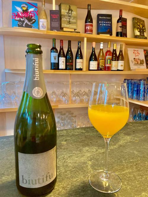 A bottle of "biutiful" sparkling wine next to a glass of orange drink, with shelves of wine bottles in the background.