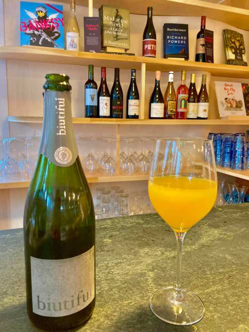 A bottle of "biutiful" sparkling wine next to a glass of orange drink, with shelves of wine bottles in the background.