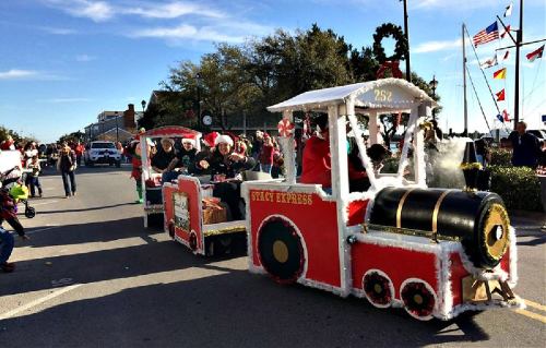 A festive train float decorated for the holidays, with people in Santa hats, parading down a street.