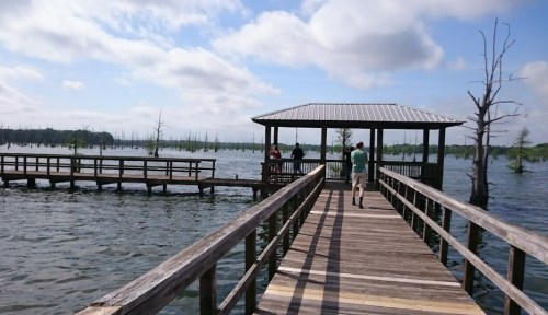 A wooden pier extends over a calm lake, with people standing in a gazebo and trees rising from the water in the background.
