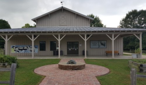 A wooden building with a metal roof, surrounded by grass and trees, featuring a central brick pathway and a sculpture.
