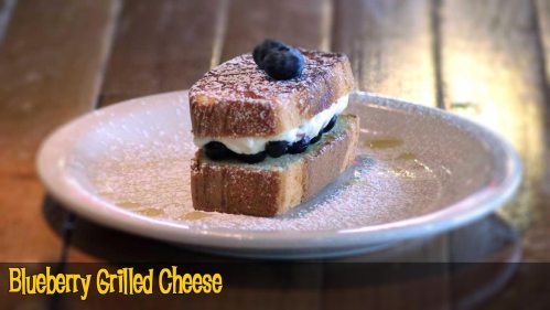 A slice of blueberry grilled cheese on a plate, dusted with powdered sugar and drizzled with syrup.