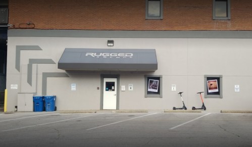 Exterior of a building with a sign reading "Rugged," featuring two scooters and framed images on the walls.