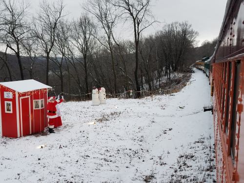 A snowy scene with a red shed, snowmen, and Santa waving near a train on a winter landscape.