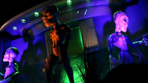Three alien figures in a colorful, illuminated setting, with green and purple lighting creating a mysterious atmosphere.