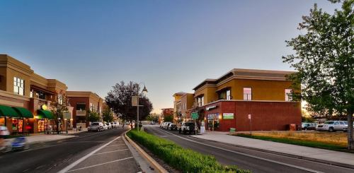 A street view of a shopping area at sunset, featuring storefronts and parked cars along a tree-lined road.