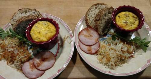 Two plates with baked egg cups, sliced ham, hash browns, and sesame bread on a wooden surface.