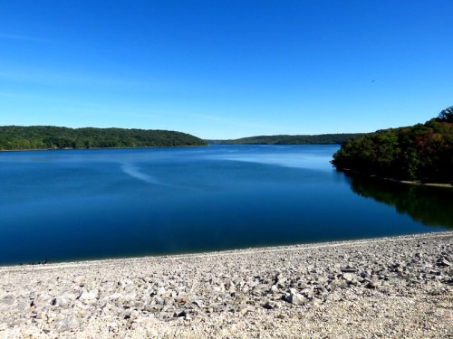 A serene lake surrounded by green hills under a clear blue sky, with a rocky shoreline in the foreground.