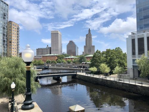 City skyline with modern buildings, a river, and a bridge under a partly cloudy sky. Lush greenery along the water.