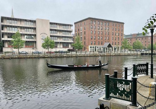 A gondola glides through a calm river, flanked by buildings and trees on a foggy day.
