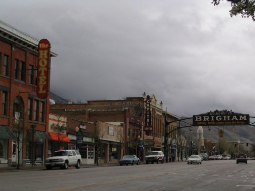 A street view of Brigham City featuring historic buildings, a hotel sign, and a welcome arch under a cloudy sky.