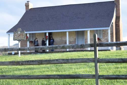 A historic house with a porch, surrounded by green grass and a wooden fence, with people standing on the porch.