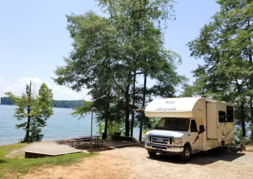 An RV parked by a serene lake, surrounded by lush green trees and a picnic area.