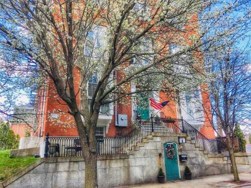 A brick building with large windows, surrounded by blooming trees and a flag hanging on the front porch.