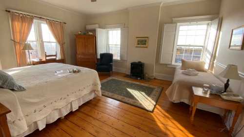 Cozy bedroom with a large bed, a small bed, a chair, and windows letting in natural light. Warm wooden floors.