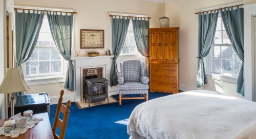 Cozy bedroom with blue carpet, a fireplace, a plaid chair, wooden dresser, and light curtains.