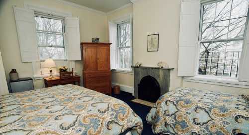 Cozy bedroom with two beds, a wooden wardrobe, and a fireplace, featuring large windows and soft lighting.