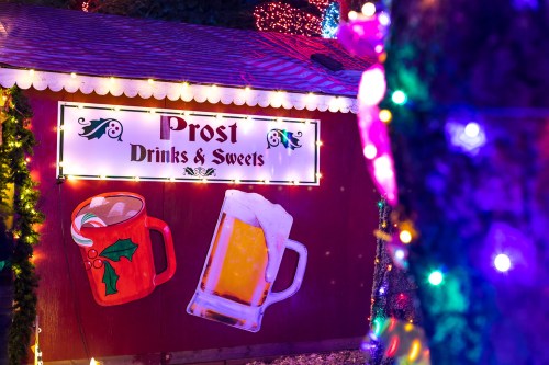 A festive drink stand with a sign reading "Prost Drinks & Sweets," decorated with holiday lights and themed mugs.