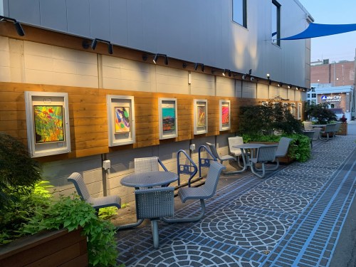 Outdoor area with framed artwork on a wooden wall, surrounded by plants and seating arrangements.