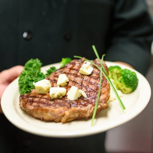A chef holds a plate with a grilled steak topped with butter and garnished with broccoli and greens.